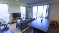 This single-floor space showcases a thoughtfully designed open-concept layout that seamlessly connects the living, dining, and kitchen areas. Large floor-to-ceiling windows dominate the exterior walls, flooding the interior with abundant natural light while offering expansive views of the surrounding cityscape. The living area is anchored by a sleek, low-profile sofa in a cool blue-grey tone, complemented by a minimalist media console housing a large flat-screen television. Track lighting along the ceiling provides adjustable ambient illumination throughout the space. The dining area features a substantial dark-toned table with seating for multiple guests, positioned to take full advantage of the panoramic window views. A smaller secondary dining or breakfast nook is visible near the corner windows, ideal for casual meals. Decorative elements, including tall dried branches in a floor vase and a motivational wall sign, add personality to the neutral-toned walls. The overall ambiance is contemporary and airy, characterized by clean lines, a cohesive colour palette of greys and warm neutrals, and an emphasis on maximizing natural light and urban views throughout the living space.