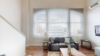 Located at 50 N Illinois St in the heart of downtown Indianapolis, this two-floor urban loft offers a striking blend of modern design and city living. The main living area commands attention with dramatically soaring ceilings and a wall of large arched windows fitted with horizontal blinds, flooding the space with natural light and framing sweeping views of the Indianapolis skyline. Rich hardwood floors anchor the open-concept living and dining area, creating a warm contrast against the bright, neutral walls.

The kitchen is well-appointed with light wood cabinetry, granite countertops, stainless steel appliances including a refrigerator, electric range, and built-in microwave, complemented by a tiled backsplash and a contemporary pendant light fixture overhead.

The bedroom is generously sized with plush carpeting, exposed structural elements on the ceiling, a large closet with wood-panel doors, and a window that allows natural light to filter in softly. An en-suite bathroom connects directly to the bedroom and features a vanity with granite-style countertops and wood cabinetry, while a second bathroom includes a shower-tub combination. The two-floor layout, visible in the architectural overview, adds dimension and versatility to this dynamic downtown residence.