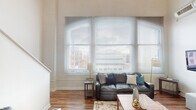 Located at 50 N Illinois St in the heart of downtown Indianapolis, this two-floor urban loft offers a striking blend of modern design and city living. The main living area commands attention with dramatically soaring ceilings and a wall of large arched windows fitted with horizontal blinds, flooding the space with natural light and framing sweeping views of the Indianapolis skyline. Rich hardwood floors anchor the open-concept living and dining area, creating a warm contrast against the bright, neutral walls.

The kitchen is well-appointed with light wood cabinetry, granite countertops, stainless steel appliances including a refrigerator, electric range, and built-in microwave, complemented by a tiled backsplash and a contemporary pendant light fixture overhead.

The bedroom is generously sized with plush carpeting, exposed structural elements on the ceiling, a large closet with wood-panel doors, and a window that allows natural light to filter in softly. An en-suite bathroom connects directly to the bedroom and features a vanity with granite-style countertops and wood cabinetry, while a second bathroom includes a shower-tub combination. The two-floor layout, visible in the architectural overview, adds dimension and versatility to this dynamic downtown residence.