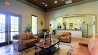 This single-story space is designed with an open, flowing layout that seamlessly connects a comfortable lounge area to a counter-style kitchen or service nook. The lounge is anchored by rich brown leather seating, including a full sofa and a matching armchair, arranged around a dark wood coffee table with ornate carved legs, all set atop a decorative area rug. Crown molding and a contrasting dark-painted tray ceiling add architectural character, while recessed lighting provides warm, even illumination throughout. An arched pass-through opening connects the main lounge to the adjacent kitchen or reception counter area, where pendant lights hang above the counter and a microwave and other appliances are visible. Large windows along one wall allow natural light to filter into the space, complemented by window blinds for light control. Decorative wall accents, potted greenery, and candle pillars contribute to the warm, traditional aesthetic. A small dining or bistro-style table and chairs occupy one corner, adding a functional secondary gathering spot. Overall, the space conveys a classic, inviting character suited for communal or hospitality use.