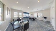 This single-floor community amenity space is designed as a stylish recreational lounge, combining comfort and entertainment in a well-appointed layout. The room is anchored by a full-size pool table positioned centrally, providing a focal point for social activity and leisure. Patterned carpet in muted blue and grey tones extends throughout, adding texture and visual interest to the floor.

The space is brightened by multiple windows along the perimeter walls, allowing generous natural light to filter in while offering views of the surrounding community. Recessed lighting in the white ceiling supplements the natural light, ensuring the room is well-lit at all times.

Seating is distributed thoughtfully across the space. Glass-top round dining tables paired with sleek black chairs provide casual gathering spots, while a dedicated bar-height table and stools are set against a bold navy blue accent wall — a striking design element that adds depth and a contemporary edge to the otherwise neutral palette. A wall-mounted flat-screen television is positioned near the entry doors, enhancing the entertainment offerings.

Framed artwork adorns the walls, contributing to the polished, modern aesthetic. A chandelier-style pendant light above the bar area introduces an elegant finishing touch. The room also includes a frosted glass interior door leading to an adjacent space, rounding out this versatile and inviting community retreat.