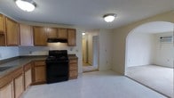 This single-floor residence offers a practical and comfortable layout designed for easy living. The kitchen is outfitted with warm honey-toned wood cabinetry providing ample storage, dark countertops, and a black electric range with an overhead ventilation hood. Light tile flooring runs through the kitchen and dining area, creating a clean and easy-to-maintain surface. A distinctive arched doorway connects the kitchen area to the adjacent living space, adding an elegant architectural touch that gives the home a sense of character and charm. The transition between rooms flows naturally, with the open sightlines making the overall floor plan feel connected and spacious. Neutral, light-toned walls throughout contribute to a bright and airy ambiance, while a hallway visible from the kitchen leads toward additional rooms, hinting at a well-organized layout. The textured ceiling adds a classic finishing detail consistent across the living areas. Overall, the space presents a welcoming and functional environment suited for everyday living, with thoughtful architectural elements that elevate its interior design beyond the purely utilitarian.