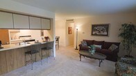 This single-floor residence offers an open, flowing layout that seamlessly connects the living, dining, and kitchen areas. The living room is anchored by plush wall-to-wall carpeting and features a comfortable seating arrangement centered around an oval coffee table, with framed artwork adding a decorative touch to the neutral-toned walls. A tall floor lamp provides warm accent lighting, enhancing the cozy atmosphere.

Adjacent to the living area, the kitchen is accessible via a breakfast bar with seating for two, making it a functional and social space ideal for casual dining. Upper cabinetry and countertop appliances are visible, offering practical storage and workspace. A passageway from the living room leads further into the residence, suggesting additional rooms beyond what is immediately visible.

The overall color palette is neutral and warm, with beige walls and light carpeting throughout, contributing to a bright and welcoming environment. The open-concept design maximizes the sense of space, while the layout promotes easy movement between living and dining functions, making it a practical and comfortable single-level home.