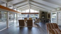 This single-floor space makes an immediate impression with its soaring vaulted ceiling, defined by clean white-painted exposed beams that draw the eye upward and lend an airy, open feel to the interior. A continuous ridge beam runs the length of the ceiling, anchoring the architectural symmetry and adding warmth to the overall design.

The living area is generously proportioned and bathed in natural light, courtesy of a dramatic wall of large, triangular and rectangular windows that frame views of the surrounding outdoor deck and neighborhood beyond. A sliding glass door provides seamless access to the expansive deck, blurring the boundary between indoor and outdoor living.

Dark hardwood flooring runs throughout, offering a rich contrast to the bright white ceiling and light-toned walls. The living space is arranged around a patterned area rug, anchoring a comfortable seating arrangement with upholstered armchairs in warm amber tones. A dining area with a table and chairs is positioned nearby, maximizing the open layout.

Track lighting mounted along the ceiling beams ensures versatile and well-distributed illumination throughout the space. A decorative freestanding shelving unit adds functional character to one corner. Overall, the space combines mid-century architectural charm with an inviting, well-lit ambiance ideal for both relaxing and entertaining.