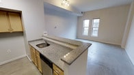 This single-floor space showcases a thoughtfully designed open-concept layout that seamlessly connects the kitchen and living areas. The kitchen is a standout feature, centered around a generous granite-topped peninsula with a speckled grey finish that provides ample prep and bar-style seating space. It is equipped with warm honey-toned cabinetry, a stainless steel undermount sink, and a stainless steel dishwasher. Overhead, a modern track lighting fixture illuminates the workspace effectively. The kitchen flows naturally into the living area, which benefits from polished concrete floors that lend an industrial yet clean aesthetic throughout the space. Two large windows in the living area allow generous natural light to fill the room, offering views of the surrounding exterior. The ceilings are notably elevated, contributing to an open and airy atmosphere. The neutral white walls throughout provide a versatile backdrop, making the space highly adaptable for a variety of interior styles. The overall ambiance is modern and functional, with a cohesive design language that balances practicality with contemporary aesthetics.