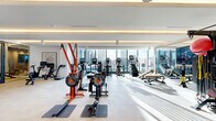 Located at 3130 Potomac Ave in Arlington, Virginia, this upscale fitness center offers a thoughtfully designed, single-level space that blends functionality with contemporary aesthetics. The open layout is bathed in natural light from expansive floor-to-ceiling windows, which frame sweeping views of the surrounding Arlington skyline and create an energizing workout environment.

The facility is organized into distinct zones catering to a wide range of fitness activities. The main cardio and strength area features a comprehensive selection of equipment, including rowing machines, stationary bikes, stair climbers, treadmills, cable machines, and weight benches, all arranged to maximize space and flow. Striking orange-accented equipment adds a bold visual element against the clean white walls and light-toned tile flooring.

A dedicated free weights and functional training zone is defined by an elegant vertical slatted gold room divider, lending a refined architectural touch. This area is stocked with kettlebells, medicine balls, resistance accessories, and TRX systems. A separate enclosed studio with glass walls and light wood flooring provides a private space for cycling or focused training sessions, equipped with its own mirror wall for form feedback.

Throughout the space, recessed cove lighting complements the natural light, and curated artwork on the walls contributes to a polished, upscale atmosphere. A lounge seating area with a plush armchair offers a comfortable retreat within the facility.