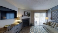This single-floor space offers a well-appointed and inviting layout designed for comfortable everyday living. The living area serves as the focal point of the home, anchored by a tile-surround fireplace with a clean white mantel — an ideal spot for decorative accents and a warm, welcoming ambiance. Dark hardwood-style flooring runs throughout, providing a rich contrast against the neutral, light gray walls and textured ceiling. A large sliding glass door fitted with vertical blinds floods the room with natural light while offering direct access to an outdoor patio, creating a seamless connection between interior and exterior spaces. The living area is generously sized, comfortably accommodating a seating arrangement alongside a media console and wall-mounted television. The overall aesthetic blends modern and rustic elements, with wood-toned cabinetry and soft, neutral tones throughout. The space is thoughtfully designed to maximize both functionality and comfort, making it an ideal environment for relaxing or entertaining.