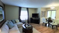 This single-floor residence offers a comfortable and thoughtfully designed living environment. The main living area is anchored by warm hardwood-style flooring that runs throughout, providing a cohesive and inviting atmosphere. Neutral sage-green walls complement the light-toned sectional sofa, which is arranged around a dark, oval coffee table atop a plush area rug — creating a relaxed and functional seating arrangement. A media console with a flat-screen television serves as the focal point of the room, flanked by decorative accents including a botanical vase arrangement and wall art that add character to the space. A bright window dressed with dark curtains and horizontal blinds allows natural light to filter into the room, enhancing the open feel. The textured ceiling adds a classic touch to the interior finish. Just beyond the living area, a glimpse of the dining space is visible, featuring white dining chairs that echo the clean, modern aesthetic carried throughout the home. The overall ambiance is warm, approachable, and well-balanced, making it an ideal space for both relaxation and everyday living.