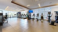 Located at 1180 Rue de Bleury in the heart of Montréal, this fitness facility offers a generously sized, single-floor exercise room designed to accommodate a comprehensive range of workout activities. The space features a refined interior aesthetic, with a coffered ceiling accented by warm ambient LED lighting along tiered crown moldings, complemented by recessed pot lights and large-format neutral floor tiles throughout.

The exercise room is divided into distinct functional zones. The cardio area is lined with treadmills, stationary bikes, rowing machines, and elliptical trainers, all positioned to take advantage of expansive floor-to-ceiling windows that flood the space with natural light and provide open views of the surrounding Montréal cityscape. Mounted flat-screen televisions are positioned for easy viewing during workouts.

The strength training area is equally well-appointed, featuring a full rack of dumbbells, adjustable benches, cable machines, and a variety of plate-loaded and selectorized resistance machines. A dedicated free weights zone includes squat racks and barbells. Full-length mirrors along the walls enhance the sense of openness and allow for form monitoring during exercise.

Warm wood-toned accent panels and wall sconces add a polished, upscale character to the space, making this gym both functional and visually inviting.