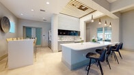 Located at 2600 Executive Drive in Venice, Florida, this single-floor community amenity space offers a sophisticated and welcoming environment designed for both social gatherings and everyday relaxation. The layout flows seamlessly from zone to zone, unified by large-format cream tile flooring that runs throughout the entire space.

The grand lounge area commands immediate attention with its soaring coffered ceilings, dramatic sputnik-style chandeliers, and floor-to-ceiling windows that frame views of palm trees and the surrounding community. Generous seating arrangements, including plush armchairs and sofas, create an inviting atmosphere bathed in abundant natural light.

The kitchen and bar area is a standout feature, anchored by a curved white quartz island with bar seating and complemented by a mosaic tile backsplash, built-in cabinetry, and a wall-mounted television. A distinctive slatted wood ceiling detail above the kitchen adds warmth and architectural interest. Pendant lighting with teardrop glass fixtures enhances the contemporary aesthetic.

Adjacent dining areas offer multiple seating configurations, from communal tables to intimate settings, all accented by soft blue-gray wall tones and curated artwork. Large windows at the far end of the dining space flood the room with natural light, creating a bright and airy ambiance. Sliding frosted glass doors provide access to additional private areas within the facility.