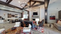 Located at 3130 Hillandale Rd in Durham, NC, this single-floor clubhouse facility delivers a sophisticated blend of rustic warmth and contemporary design. The entrance foyer sets an impressive tone with soaring vaulted ceilings, herringbone-patterned tile flooring, and a statement round chandelier with exposed Edison bulbs, flanked by floor-to-ceiling glass French doors that flood the space with natural light.

The expansive lounge area is a standout, featuring dramatic dark wood ceiling beams and white shiplap detailing overhead. A floor-to-ceiling stacked stone fireplace anchors the room, paired with a wall-mounted television and a reclaimed wood mantel. Seating is arranged around a bold, abstract-patterned area rug in deep reds and grays, complemented by mixed-material furnishings that balance comfort and style.

Adjacent to the lounge, the dining and kitchen area offers a large communal table beneath a linear glass pendant light. The kitchen features crisp white cabinetry, sliding barn doors in weathered gray, and stainless steel appliances—ideal for gatherings and events. Dark wood accent walls and reclaimed wood paneling appear throughout, unifying the overall aesthetic.

Additional seating nooks and open corridors with abundant natural light create a welcoming, versatile environment well-suited for both relaxation and social activities.