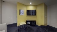 This single-floor game zone is purpose-built for interactive entertainment, combining comfortable furnishings with a thoughtfully curated gaming atmosphere. The main lounge area features a spacious light grey sectional sofa positioned beneath two framed watercolor-style prints of game controllers, lending the space a fun yet polished aesthetic against the textured neutral walls.

An adjacent gaming area is defined by bold yellow accent walls and houses a wall-mounted flat-screen television paired with a low-profile black media console storing gaming consoles and accessories. A framed black-and-white print of classic game controllers adds a nostalgic touch to this zone, while dark carpeting anchors the space and a small yellow ottoman provides additional seating.

A dedicated co-op gaming nook accommodates two X Rocker-style gaming chairs positioned before a shared table surface, ideal for multiplayer sessions. A framed "Periodic Table of Controllers" decorates the wall, reinforcing the gaming theme. Dark bean bags or floor cushions are also present throughout, offering casual and flexible seating options. Recessed lighting provides consistent illumination across all areas. The space connects to an adjacent fitness area, suggesting it is part of a larger shared-amenity facility.