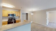 This single-floor residence offers a practical and comfortable layout designed for everyday living. The open-concept kitchen and living area form the heart of the home, where light wood-toned cabinetry, granite-style countertops, and a full suite of stainless steel appliances — including a refrigerator, electric range, built-in microwave, and dishwasher — are complemented by easy-care hardwood-style flooring. A curved breakfast bar peninsula provides additional prep space and casual seating, while a pendant light above adds a decorative touch. The adjacent living area is finished with plush neutral carpet and flows seamlessly through the space, with a tiled entry foyer visible from the main corridor. Two bedrooms feature ceiling fans with light fixtures, large windows with vertical blinds for natural light and privacy, and generous walk-in closets with built-in shelving. Both bathrooms are similarly appointed, each offering a full tub-shower combination with white tile surround, a wood-toned vanity with an integrated sink, and rich dark slate-style tile flooring. Additional storage is provided by open shelving visible in the living area. The neutral color palette throughout creates a calm, cohesive ambiance well-suited to a variety of furnishing styles.