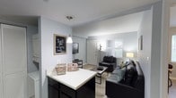 This single-floor unit at Campus Court at Red Mile presents an open-concept design that seamlessly connects the kitchen and living areas. The kitchen features dark cabinetry with a white countertop that extends into a breakfast bar, providing both additional workspace and casual seating. A pendant light hangs above the counter, adding a focused lighting element to the space.

The living area is generously proportioned, accommodating a full seating arrangement including a leather sofa and accent chair centered around a coffee table. A wall-mounted television and media console provide an entertainment focal point, while a floor lamp adds ambient lighting. The room benefits from natural light through a window that also offers a view of the surrounding greenery outside.

The flooring transitions from the kitchen area into the living space, which features carpet, adding comfort underfoot. The walls are painted in a soft, neutral tone throughout, contributing to a clean and cohesive aesthetic. The layout is efficient and practical, making it well-suited for everyday living in a community setting near campus.
