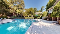Located at 100 Chattahoochee Circle in Roswell, Georgia, this outdoor amenity space centers around a generously sized swimming pool with crisp blue water and tiled edges. The pool deck is finished in light-toned stone pavers, providing ample room for lounging and entertaining. A standout feature is the wood pergola structure positioned alongside the pool, sheltering a stone-faced bar area — ideal for outdoor gatherings. Multiple lounge chairs and umbrella-shaded seating are arranged along the pool's perimeter, offering both sun and shade options for guests. The surrounding landscape is meticulously maintained, with neatly trimmed topiary hedges, sculpted shrubs, and a variety of mature trees that create a lush, private atmosphere. A staircase with white railings leads to an elevated area, adding a layered architectural dimension to the outdoor environment. The mature tree canopy frames the space, filtering natural sunlight and enhancing the sense of seclusion. The overall ambiance is refined and resort-like, blending classic landscaping with functional outdoor amenities in a well-appointed setting characteristic of Roswell's established residential communities.