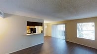 This single-floor space offers a well-connected open layout that seamlessly blends the kitchen and living areas. The kitchen features a raised pass-through counter with dark cabinetry overhead, providing both a functional workspace and a natural divide between the cooking and living zones. A black refrigerator and sink are visible within the kitchen, complemented by warm wood-toned upper cabinets that add a rich contrast to the light-colored walls.

The living area is generously proportioned, showcasing rich, dark hardwood-style flooring that runs throughout and adds a grounding warmth to the space. A textured popcorn ceiling spans the entirety of the unit, adding a classic architectural character. A decorative pendant light fixture serves as a subtle focal point overhead, providing ambient illumination to the open space.

One of the standout features of the living area is the full-width sliding glass door fitted with vertical blinds, which floods the interior with abundant natural light and offers direct access to the outdoors. The clean, neutral wall tones throughout create a versatile backdrop, making the space easy to personalize. Overall, the layout and features combine to create a functional, light-filled environment well-suited for everyday living.