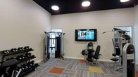 This single-floor fitness space is thoughtfully designed to accommodate a range of workout routines within a well-organized layout. The room features a dark-painted ceiling that contrasts with the light-toned walls, creating a focused and energetic atmosphere. Bright circular overhead lights illuminate the space effectively, ensuring excellent visibility throughout.

The flooring consists of multi-tone carpet tiles in grey, orange, and yellow, adding a modern, dynamic visual element while providing cushioning underfoot. A wall-mounted flat-screen television is positioned prominently, allowing users to stay entertained or follow workout programs during their sessions.

The equipment selection is practical and varied. A multi-functional cable machine anchors one side of the room, suitable for a wide range of strength-training exercises. A dumbbell rack stocked with multiple weight increments sits nearby, catering to free-weight training. An adjustable weight bench paired with a plate-loaded machine rounds out the strength equipment. A mirrored entry or adjoining area at the rear enhances the sense of depth and allows users to monitor their form. Overall, this fitness space offers a clean, functional environment for effective daily workouts.