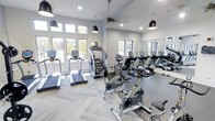 This fitness center offers a bright, welcoming environment designed to support an active lifestyle. The space is bathed in natural light streaming through a generous array of large windows along multiple walls, providing pleasant outdoor views of surrounding greenery and keeping the atmosphere open and energizing. Elevated ceilings with a ceiling fan enhance airflow and comfort during workouts, while stylish pendant lighting adds a modern touch to the overall aesthetic. The flooring features a neutral, textured carpet tile in a herringbone pattern, lending both visual appeal and practical underfoot support. A full-length wall mirror expands the sense of space and allows for easy form-checking during exercise. The cardio equipment lineup is comprehensive and well-maintained, including treadmills, stationary bikes, and a stair climber, all from modern fitness brands. The layout is thoughtfully arranged to provide ample space between machines, ensuring a comfortable and unobstructed workout experience. The neutral gray color palette and clean, contemporary finishes create a professional yet approachable atmosphere, making this fitness center a functional and visually appealing amenity.