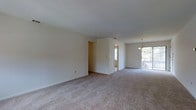This single-floor space presents an open, understated layout finished in neutral cream tones throughout. The main living area is generously proportioned, featuring wall-to-wall carpet in a light beige hue that contributes to the overall warmth of the interior. Smooth, white ceilings and clean white baseboards frame the space, lending a simple yet tidy aesthetic.

A partial interior wall creates a subtle visual division within the open plan, defining separate zones while maintaining an airy, connected feel. Natural light filters in from multiple directions, including a window visible in the background and sliding glass doors at the far end of the space that open to what appears to be an outdoor patio or balcony area, offering views of surrounding greenery.

A chandelier with a classic design is suspended near the dining or transitional area, adding a modest decorative element. The sliding glass doors allow ample daylight to fill the space, enhancing its bright and open character. Overall, the space offers a clean, functional environment with a neutral palette that provides a versatile backdrop for a variety of uses.
