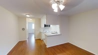 This single-floor space offers a practical and comfortable layout centered around an open-concept living and kitchen area. The kitchen is designed with white cabinetry, white appliances including a range, and an over-the-range microwave. A built-in peninsula with an integrated sink provides additional counter space and serves as a natural divider between the kitchen and the adjoining living area, creating a semi-open flow throughout.

The living area features warm hardwood-style flooring that extends throughout the unit, lending a cohesive and inviting atmosphere. A ceiling fan with a light fixture is centrally positioned, providing both air circulation and general illumination. The walls are finished in a neutral off-white tone, contributing to a clean and bright interior.

A hallway visible from the living area connects to additional rooms, with natural light streaming in through a window at the far end, enhancing the overall sense of openness. Flush-mount lighting fixtures are installed throughout the hallway, ensuring consistent illumination. The unit's layout is straightforward and efficiently designed, making good use of the available space while maintaining a welcoming, functional environment.