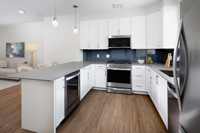 Phase II Renovated Package I townhome kitchen with stainless steel appliances, grey quartz countertops, white cabinetry, grey tile backsplash, and hard surface flooring - Avalon Acton