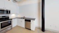 This 1-bedroom, 1-bathroom loft-style unit showcases a compelling blend of industrial character and modern finishes across a single open floor. Polished concrete floors run throughout the space, complementing the exposed ductwork and structural elements that lend an authentic urban loft aesthetic.

The kitchen is well-appointed with crisp white cabinetry extending to ceiling height, dark granite countertops, and a full suite of stainless steel appliances including a refrigerator, electric range, over-the-range microwave, and dishwasher. White subway tile backsplash adds a clean, classic contrast to the dark counters, while a stainless steel sink anchors the functional layout.

The living area is generously proportioned with soaring ceilings that enhance the sense of openness, exposed wood ceiling details above, and prominent structural columns that reinforce the building's industrial heritage. The bedroom offers a quiet, neutral retreat with the same concrete flooring and continued exposure to the architectural ductwork above.

The bathroom is clean and functional, featuring tile flooring in a light wood-look pattern, a white vanity with granite countertop, an undermount sink, and a full tub and shower combination. Recessed lighting throughout provides focused illumination, while the overall white and neutral palette keeps the space feeling bright and airy.