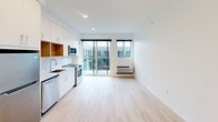 This efficiently designed studio offers a thoughtfully organized single-floor layout that maximizes every square foot. Light wood flooring runs continuously throughout, lending a clean and cohesive aesthetic to the entire space.

The open living area is bright and airy, illuminated by recessed ceiling lighting and generous natural light from a floor-to-ceiling sliding glass door that opens onto a private balcony. The outdoor view provides an urban backdrop, enhancing the sense of openness within the unit.

The kitchen is well-appointed and modern, featuring white flat-panel cabinetry with warm wood open shelving accents, a classic white subway tile backsplash, and sleek light quartz countertops. It is fully equipped with stainless steel appliances, including a refrigerator, dishwasher, built-in microwave, and a range — offering full functionality in a compact footprint.

The bathroom is clean and minimalist in design, with white cabinetry, a single-basin vanity, and a full soaking tub with shower. Brushed nickel fixtures and light wood flooring carry the modern theme throughout. The all-white palette creates a bright and fresh atmosphere. Overall, the space presents a contemporary and functional living environment with a neutral, move-in-ready finish.