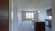 This single-floor space showcases an open-concept layout that seamlessly connects the kitchen and living areas. The kitchen peninsula is a standout feature, topped with light granite countertops and supported by rich, dark-stained cabinetry with sleek hardware. Two elegant pendant lights hang above the peninsula, providing focused task lighting, while a track lighting system runs along the ceiling to illuminate the broader space.

The living area extends naturally from the kitchen, finished with plush, neutral-toned carpeting that adds warmth and comfort underfoot. Smooth white walls and clean white baseboards contribute to a bright, airy aesthetic throughout. A large multi-pane window at the far end of the living area invites generous amounts of natural light and offers views of the surrounding neighborhood and greenery, enhancing the open feel of the space.

A frosted glass door near the entrance adds a subtle architectural detail while maintaining privacy. The overall ambiance is clean, modern, and functional, with a neutral palette that provides a versatile backdrop adaptable to a variety of interior design preferences.