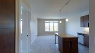This single-floor space showcases an open-concept layout that seamlessly connects the kitchen and living areas. The kitchen peninsula is a standout feature, topped with light granite countertops and supported by rich, dark-stained cabinetry with sleek hardware. Two elegant pendant lights hang above the peninsula, providing focused task lighting, while a track lighting system runs along the ceiling to illuminate the broader space.

The living area extends naturally from the kitchen, finished with plush, neutral-toned carpeting that adds warmth and comfort underfoot. Smooth white walls and clean white baseboards contribute to a bright, airy aesthetic throughout. A large multi-pane window at the far end of the living area invites generous amounts of natural light and offers views of the surrounding neighborhood and greenery, enhancing the open feel of the space.

A frosted glass door near the entrance adds a subtle architectural detail while maintaining privacy. The overall ambiance is clean, modern, and functional, with a neutral palette that provides a versatile backdrop adaptable to a variety of interior design preferences.