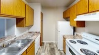 This single-floor residential unit offers a well-organized layout designed for practical, comfortable living. The kitchen is a standout functional space, featuring warm honey-toned oak cabinetry lining both walls, laminate countertops with a stone-like finish, a double-basin sink, a white electric range, and a full-size refrigerator. Wood-look vinyl plank flooring runs throughout the kitchen, adding a warm, durable finish underfoot.

The living area flows naturally from the kitchen through an open pass-through design, with plush carpet underfoot and neutral gray walls that create a calm, versatile backdrop. A front entry door opens directly into this space, providing easy access and a clear sense of arrival.

The unit includes multiple carpeted bedrooms with neutral tones and clean white ceilings. At least one bedroom benefits from a window with blinds, allowing natural light to brighten the space. The rooms offer ample wall space suitable for various furniture arrangements.

Two bathrooms serve the unit, each featuring wood-look vinyl plank flooring that echoes the kitchen's aesthetic. One bathroom includes a vanity with a white countertop and warm wood cabinetry, a mirror, and a toilet. The second bathroom is more compact, with a toilet and access to an adjacent area. Both bathrooms are finished in clean, neutral tones with overhead ventilation.
