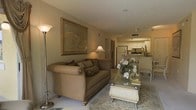 This single-floor space presents an open-concept layout that seamlessly connects the living area to the kitchen. The living room is anchored by a plush, tan upholstered sofa adorned with decorative pillows, positioned atop a patterned area rug. A glass-top coffee table with ornate detailing serves as a central focal point, complemented by a tasteful floral arrangement. Warm ambient lighting is provided by a tall floor lamp and a classic table lamp, creating a cozy atmosphere. A large framed artwork depicting a decorative urn adds a refined touch to the neutral-toned walls, which feature a smooth textured ceiling above. The open kitchen, visible in the background, includes white cabinetry, a refrigerator, and a raised breakfast bar with seating. Track lighting illuminates the kitchen workspace, while the light-toned flooring ties both spaces together cohesively. The overall ambiance is warm and traditionally styled, with classic furnishings and neutral earth tones creating an inviting and comfortable living environment throughout the space.