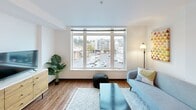 Located at 12739 Lake City Way NE in Seattle's Lake City neighborhood, this efficiently designed apartment on the second floor offers a thoughtful layout across a single level, comprising a kitchen, living room, and bathroom. The galley-style kitchen is a standout feature, outfitted with stainless steel appliances including a full-size refrigerator, electric range, built-in microwave, and dishwasher. Dark lower cabinetry is complemented by warm wood-toned upper cabinets and clean white countertops, all set atop durable wood-look flooring that runs throughout the unit. Recessed lighting illuminates the kitchen, creating a bright and functional cooking space. The living area benefits from a large, multi-pane picture window that fills the room with natural light and offers open views of the surrounding Lake City neighborhood and Seattle skyline. The clean white walls and light-toned interior contribute to an airy, modern ambiance throughout. The bathroom features a dark wood vanity with a white countertop, an integrated sink, a large mirror, and neutral-toned flooring, maintaining the unit's consistent contemporary aesthetic. The overall design emphasizes clean lines, efficient use of space, and modern finishes, making this a well-appointed urban residence in a convenient Seattle location.
