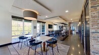 This single-floor communal space is designed to accommodate a variety of uses, blending lounge, dining, co-working, and meeting functions within a cohesive, contemporary setting. The layout flows openly from a welcoming entry corridor—flanked by a tan leather sofa and polished concrete floors—into a series of distinct yet interconnected zones.

The main gathering area features marble-topped bistro tables paired with modern side chairs, all set atop patterned area rugs that anchor the space. Oversized windows line the perimeter, flooding the interior with natural light and offering views of surrounding greenery. High bar-height seating runs along one wall, accompanied by a wall-mounted flat-screen television, making it suitable for both casual socializing and informal presentations.

A dedicated conference room provides a more private setting, centered around a long yellow-topped table with upholstered bar chairs, pendant lighting overhead, and additional wall-mounted display screens. Brick accent columns punctuate the open floor plan, adding an industrial warmth that contrasts with the otherwise clean, modern aesthetic. Open metal shelving units serve as partial dividers between zones while maintaining visual connectivity throughout. The overall ambiance is bright, functional, and polished.