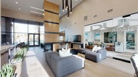 This stunning luxury lobby is a masterclass in contemporary design, immediately commanding attention with its soaring double-height ceilings and refined finishes. A dramatic floor-to-ceiling wood-clad fireplace column serves as the centerpiece of the space, adding warmth and architectural grandeur. Pendant lighting cascades elegantly from above, casting a warm, inviting glow throughout the expansive interior.

The lounge area is appointed with plush, tufted gray sectional sofas arranged around a sleek coffee table, creating a comfortable yet sophisticated gathering space. Light wood-toned flooring runs throughout, complementing the neutral palette of warm grays, whites, and natural wood tones that define the overall aesthetic.

Floor-to-ceiling glass walls and large windows flood the space with abundant natural light while offering views of the surrounding exterior, seamlessly blending the indoors with the outside environment. A wall-mounted flat-screen TV is integrated into a clean-lined media wall, adding a functional element without disrupting the design flow.

The overall ambiance is polished and modern, with every detail thoughtfully curated to convey luxury and comfort. This space sets a high standard, reflecting the elevated lifestyle associated with the DUO Luxury brand prominently featured throughout.