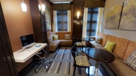 This thoughtfully designed hotel suite sitting room combines classic sophistication with modern functionality. The interior is defined by rich, dark wood-paneled walls that lend a warm, intimate ambiance throughout the space. Two windows fitted with horizontal blinds and decorative valances allow natural light to filter in while maintaining privacy, framing the room with an elegant touch.

A wraparound upholstered sectional sofa lines the corner beneath the windows, accented with patterned and solid throw pillows in earthy tones of gold, green, and orange. Adjacent to the sofa sits a small round dark wood dining table paired with coordinating upholstered chairs featuring a bold geometric pattern — creating a cozy spot for casual dining or conversation.

On the opposite side of the room, a dedicated workspace features a sleek desk with an ergonomic chair, complete with a computer monitor, making it well-suited for business travelers. Wall-mounted sconce lighting adds a refined, hotel-classic aesthetic, complementing the overall warm color palette.

The floor is covered with a multi-toned striped area rug in shades of gray, blue, and beige, adding texture and visual interest to the room. A framed map print adorns one wall, contributing a subtle, travel-inspired decorative element. The overall design balances comfort, utility, and classic style.