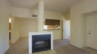 This single-floor residence offers an open-concept layout that promotes a sense of spaciousness and connectivity throughout the living areas. The main living space is anchored by a striking freestanding fireplace with a clean white surround and tiled hearth, serving as a natural focal point that divides and defines the room. A cylindrical support column rises alongside it, adding an architectural element that contributes to the space's distinct character.

The living area features neutral-toned carpeting that extends throughout, creating a warm and cohesive foundation underfoot. Smooth, light-colored walls and standard ceiling heights contribute to a bright, understated ambiance, with ceiling vents ensuring consistent climate control.

Flowing seamlessly from the living area, the kitchen is partially partitioned by a half-wall counter that provides both workspace and a visual connection to the main room. The kitchen is equipped with dark wood upper cabinetry contrasted against lighter lower units, a functional sink, and white appliances. The overall palette of the space — neutral walls, warm wood tones, and soft carpeting — creates a simple and livable environment suited for everyday comfort.
