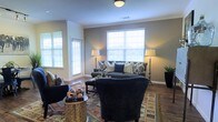 This single-story space showcases a thoughtfully designed living area that balances comfort and style. The sitting room serves as the focal point, featuring warm-toned walls accented by crisp white crown molding and baseboards that lend a polished, finished look throughout. Large windows fitted with white plantation shutters allow generous amounts of natural light to fill the room while offering privacy and light control.

The hardwood flooring grounds the space and complements the layered area rug beneath the seating arrangement, which includes a dark upholstered sofa adorned with an assortment of patterned throw pillows and a matching accent chair. Tall floor lamps on either side of the sofa provide warm ambient lighting, enhancing the inviting atmosphere. A rectangular coffee table and a decorative side table complete the arrangement, offering both functionality and visual interest.

The layout allows for easy flow between areas, with a doorway visible to one side suggesting additional rooms beyond. A potted plant adds a touch of greenery, contributing to the overall warm and welcoming ambiance of this well-maintained space.