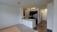 This single-floor residence features an open-concept layout that seamlessly connects the kitchen and living area. The kitchen is outfitted with dark espresso cabinetry that provides a striking contrast against the white peninsula base, which is topped with a light granite-style countertop. The peninsula includes a built-in sink and offers additional prep space while also serving as a natural divider between the kitchen and the carpeted living area. Stainless steel refrigerator and black appliances, including a range and microwave, complete the kitchen's functional design. Track lighting overhead illuminates the cooking space efficiently. The flooring transitions from wood-style planks in the kitchen to plush carpet in the living area, adding warmth and texture. The walls are finished in a clean, neutral white, contributing to a bright and airy atmosphere. A hallway visible in the background leads to additional rooms, suggesting a thoughtful and functional floor plan. Ceiling vents indicate a central HVAC system throughout the unit.