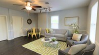 This single-floor space offers a welcoming open-concept layout that seamlessly connects the living and dining areas into one cohesive, functional environment. Dark hardwood flooring runs throughout, providing a rich contrast to the soft gray walls accented by classic chair-rail wainscoting. The living area is anchored by a light gray sofa and armchair arrangement centered around a round white coffee table, all set atop a bold green and cream geometric area rug that adds a lively focal point to the room. Track lighting and a ceiling fan with an integrated light fixture provide versatile illumination, while a window allows natural light to brighten the space. A framed mirror on the wall enhances the sense of openness. The dining area features a dark table paired with distinctive yellow metal chairs, adding a vibrant pop of color that complements the green accents found throughout. A decorative sunburst mirror and fresh floral arrangements contribute to the space's polished, contemporary character. Overall, the design balances comfort and style through a coordinated palette of gray, yellow, and green, creating an inviting atmosphere well-suited for both everyday living and entertaining.