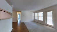 This single-floor residence offers a clean, functional layout designed for comfortable everyday living. The main living area is a standout feature, combining plush wall-to-wall carpeting with rich hardwood flooring that transitions seamlessly between spaces. Multiple windows line the exterior wall, flooding the room with natural light and providing views of the surrounding outdoor area, including an adjacent balcony or deck. A glass-paneled exterior door further enhances the connection to the outdoors while contributing additional brightness to the interior.

The living area is generously proportioned, offering ample room for a variety of furniture arrangements. The textured ceiling and crisp white walls provide a neutral, move-in-ready backdrop throughout. A hallway visible from the main living space leads to additional rooms, suggesting a well-organized floor plan. The transition between the carpeted living zone and the hardwood flooring area delineates distinct functional spaces within an open-concept framework.

Overall, the space presents a light, airy ambiance with practical finishes, including visible HVAC vents and standard wall outlets, making it a versatile and accommodating residential environment suited to a range of lifestyles.