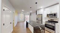 This well-appointed single-floor residence showcases a thoughtfully designed open-concept layout that seamlessly connects the kitchen and living areas. The kitchen is the undisputed centerpiece, anchored by a striking large island clad in dramatic black-and-white veined stone that extends to a waterfall edge. White shaker cabinetry lines the walls from counter to ceiling, providing generous storage, while a full suite of stainless steel appliances — including a French door refrigerator, gas range, built-in microwave, and dishwasher — offers a fully equipped cooking experience. A pair of sleek black pendant lights with gold accents hang above the island, adding a modern, sophisticated touch. Recessed lighting and light-toned hardwood flooring run throughout, enhancing the bright and airy atmosphere of the open living space. The living area benefits from natural light streaming through a window with zebra-style roller shades, alongside a glass door that opens to a private balcony with views of the surrounding neighborhood. The bathroom features a wide white vanity with marble-style countertop, a large LED-lit mirror, chrome fixtures, and clean tile flooring, delivering a polished and contemporary finish throughout.