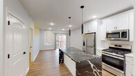 This well-appointed single-floor residence showcases a thoughtfully designed open-concept layout that seamlessly connects the kitchen and living areas. The kitchen is the undisputed centerpiece, anchored by a striking large island clad in dramatic black-and-white veined stone that extends to a waterfall edge. White shaker cabinetry lines the walls from counter to ceiling, providing generous storage, while a full suite of stainless steel appliances — including a French door refrigerator, gas range, built-in microwave, and dishwasher — offers a fully equipped cooking experience. A pair of sleek black pendant lights with gold accents hang above the island, adding a modern, sophisticated touch. Recessed lighting and light-toned hardwood flooring run throughout, enhancing the bright and airy atmosphere of the open living space. The living area benefits from natural light streaming through a window with zebra-style roller shades, alongside a glass door that opens to a private balcony with views of the surrounding neighborhood. The bathroom features a wide white vanity with marble-style countertop, a large LED-lit mirror, chrome fixtures, and clean tile flooring, delivering a polished and contemporary finish throughout.