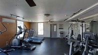 This fitness center is a dedicated, single-floor facility designed to meet a wide range of exercise needs. The space is outfitted with multiple cardio machines, including treadmills and an elliptical, positioned along one side of the room to allow for easy use and movement between equipment. On the opposite side, a multi-station strength training machine provides options for resistance and weight-based workouts. The flooring consists of dark rubber matting, which is durable, slip-resistant, and well-suited for high-activity use. The room is lit by a combination of ceiling fans with integrated lighting and track lighting mounted along the walls and ceiling, ensuring even illumination throughout the space. Two windows near the entrance allow natural light to filter in, contributing to an open and energizing atmosphere. The textured ceiling adds a classic architectural touch, while the neutral gray and accent walls create a clean, focused environment. A double-door entry provides convenient access, and a posted fitness center notice board near the entrance offers rules and guidelines for users. Overall, the space is functional, well-organized, and thoughtfully laid out to support an effective fitness routine.