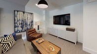This thoughtfully designed single-floor space seamlessly blends open-plan living with flexible, defined zones. The main area combines a lounge and dining space, unified by neutral carpet flooring, recessed lighting, and a muted gray-and-white color palette that creates a clean, contemporary backdrop. Large floor-to-ceiling windows dressed with teal drape panels allow generous natural light to fill the interior while framing outdoor views, contributing to an airy, welcoming atmosphere.

A standout architectural feature is the set of sliding glass barn doors with dark wood frames and black industrial hardware, which connect the main living area to an adjacent space. These doors offer the ability to open the layout for a more expansive feel or close it off for privacy, adding both function and visual character to the design.

The lounge area is anchored by a neutral sofa accessorized with bold throw pillows, alongside a rustic wood console table adorned with potted greenery and curated artwork. A dedicated media nook features a wall-mounted flat-screen television, a low white credenza, a leather lounge chair, and a warm wood coffee table, creating an intimate retreat within the broader space. Together, these zones offer a cohesive and versatile environment suited for both relaxation and gathering.