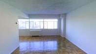 This single-floor space features a generously proportioned open living area defined by its classic parquet hardwood flooring, which reflects natural light across its polished surface. A wide bank of multi-pane windows spans much of one wall, flooding the interior with abundant daylight while framing elevated views of a surrounding urban skyline — indicative of a high-floor position within a larger building. The smooth, white walls and ceiling create a clean, neutral backdrop that complements the warm tones of the wood flooring below. A traditional radiator unit sits beneath the windows, suggesting a reliable in-unit heating system consistent with established city residential construction. The open layout of the main living area offers flexible arrangement potential, with no interior obstructions limiting usability. The overall ambiance is bright and airy, with the combination of natural light, city views, and classic architectural details like the parquet flooring lending the space a timeless urban character. The absence of interior partitions in the main area contributes to an open, uncluttered feel throughout.