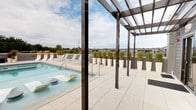 This rooftop terrace is a thoughtfully designed outdoor amenity space that combines contemporary architecture with functional leisure features. The expansive deck is finished in large-format, light-toned porcelain tiles that lend a clean, sophisticated aesthetic throughout the space.

At the heart of the terrace sits a swimming pool with clear blue water, flanked by rows of modern lounge chairs and floating pool accessories, offering an ideal setting for relaxation. A series of sleek dark-framed pergolas span portions of the deck, providing partial shade while maintaining an open, airy feel beneath the open sky.

Large planters filled with ornamental grasses are positioned along the perimeter, adding greenery and a natural element to the urban setting. An outdoor wall-mounted television is integrated into the stone-clad building facade, enhancing the entertainment possibilities of the space.

Full-height glass sliding doors and windows connect the terrace to an interior space, allowing natural light to flow freely between indoors and outdoors. Panoramic views of the surrounding cityscape, including mature treetops and neighborhood rooflines, frame the terrace on all sides, reinforcing its elevated, open-air character.