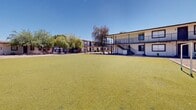 Located at 4235 N 35th Ave in Phoenix, Arizona, this multi-building property presents a well-organized campus-style layout centered around a striking, expansive courtyard finished with lush green artificial turf. The grounds are framed by a variety of mature desert and tropical trees, including towering palms and shade trees, lending the property a distinctive Southwest character beneath Phoenix's clear blue skies.

The property consists of single-story building wings arranged in a horseshoe-like configuration around the central courtyard, complemented by a two-story structure featuring an open-air walkway balcony with metal railings and staircase access. The single-story wings display a classic stucco exterior in a neutral white finish with a brown trim roofline, with multiple individual entry doors lining the facades — indicative of a multi-unit layout.

The courtyard itself serves as a prominent shared outdoor space and includes a small soccer goal, suggesting recreational use. Gravel landscaping borders portions of the buildings, requiring minimal maintenance and reflecting the practical aesthetic common to Phoenix properties. The two-story section adds vertical dimension to the property and offers covered walkway access along the ground level. Together, the buildings and grounds form a cohesive, functional property well-suited to community-oriented use in the heart of Phoenix.