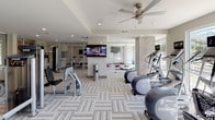 This single-level fitness center is thoughtfully designed to accommodate a wide range of workout needs within a bright, open layout. The space is divided into distinct functional zones, each outfitted with purpose-specific equipment. The cardio area features a row of treadmills and upright bikes, each equipped with individual media screens, positioned along large windows that invite generous natural light and outdoor views. Ceiling fans overhead contribute to a comfortable workout environment.

The strength training zone houses a variety of cable and resistance machines alongside free weights and benches, supported by wall-mounted televisions for entertainment. Large mirrors along the walls visually expand the space and provide functional feedback during exercise. A dedicated functional training area includes balance and agility equipment, with storage cubbies built into the base of a mirrored wall, keeping accessories organized and accessible.

The flooring throughout consists of patterned carpet tiles laid in a herringbone arrangement, adding visual texture while providing cushioning underfoot. Stone-accented columns and neutral-toned walls lend the space a clean, contemporary aesthetic. Glass entry doors connect the interior to an adjacent corridor or entryway, where additional training tools such as balance discs are stored. A water station is conveniently located within the main floor area.