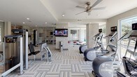 This single-level fitness center is thoughtfully designed to accommodate a wide range of workout needs within a bright, open layout. The space is divided into distinct functional zones, each outfitted with purpose-specific equipment. The cardio area features a row of treadmills and upright bikes, each equipped with individual media screens, positioned along large windows that invite generous natural light and outdoor views. Ceiling fans overhead contribute to a comfortable workout environment.

The strength training zone houses a variety of cable and resistance machines alongside free weights and benches, supported by wall-mounted televisions for entertainment. Large mirrors along the walls visually expand the space and provide functional feedback during exercise. A dedicated functional training area includes balance and agility equipment, with storage cubbies built into the base of a mirrored wall, keeping accessories organized and accessible.

The flooring throughout consists of patterned carpet tiles laid in a herringbone arrangement, adding visual texture while providing cushioning underfoot. Stone-accented columns and neutral-toned walls lend the space a clean, contemporary aesthetic. Glass entry doors connect the interior to an adjacent corridor or entryway, where additional training tools such as balance discs are stored. A water station is conveniently located within the main floor area.