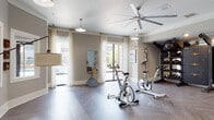 Located at 10051 Perimeter Pkwy in Charlotte, NC, this well-appointed fitness center offers a thoughtfully designed, single-floor layout that balances functionality with an inviting aesthetic. The main exercise area features herringbone-patterned hardwood-style flooring throughout, lending a polished, upscale feel to the space. Large floor-to-ceiling windows dressed with sheer white curtains flood the room with natural light and provide views of the surrounding exterior, creating an open and energizing workout environment. A large ceiling fan anchors the room overhead, complementing pendant lighting to ensure both comfort and visibility. The spin zone houses multiple stationary cycling bikes positioned to take advantage of the natural light, while a full-length wall mirror allows for form checks during exercise. A dedicated strength training station includes a Torque functional trainer, a comprehensive rack of dumbbells and kettlebells, medicine balls, and stability balls, catering to a wide range of fitness routines. The welcoming lounge area features soft blue-toned walls, a wall-mounted television, a decorative credenza, and seating for relaxation before or after a workout. Sculptural yoga-pose figurines are placed throughout, reinforcing the space's wellness-focused atmosphere. Additional amenities include closet storage and clearly posted fitness guidelines, making this facility both practical and aesthetically refined.